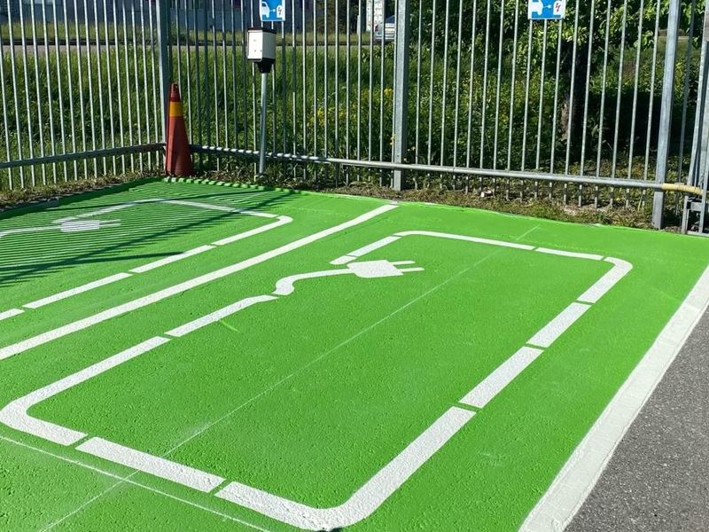 Två laddplatser med grön yta, vita kantlinjer och en symbol med kontakt och sladd, framför ett staket med laddstolpe.